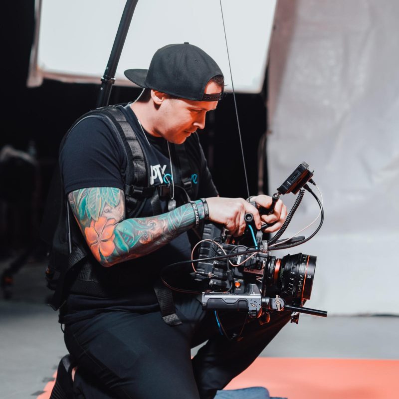 A video producer with tattoos and a cap adjusts camera equipment on set.